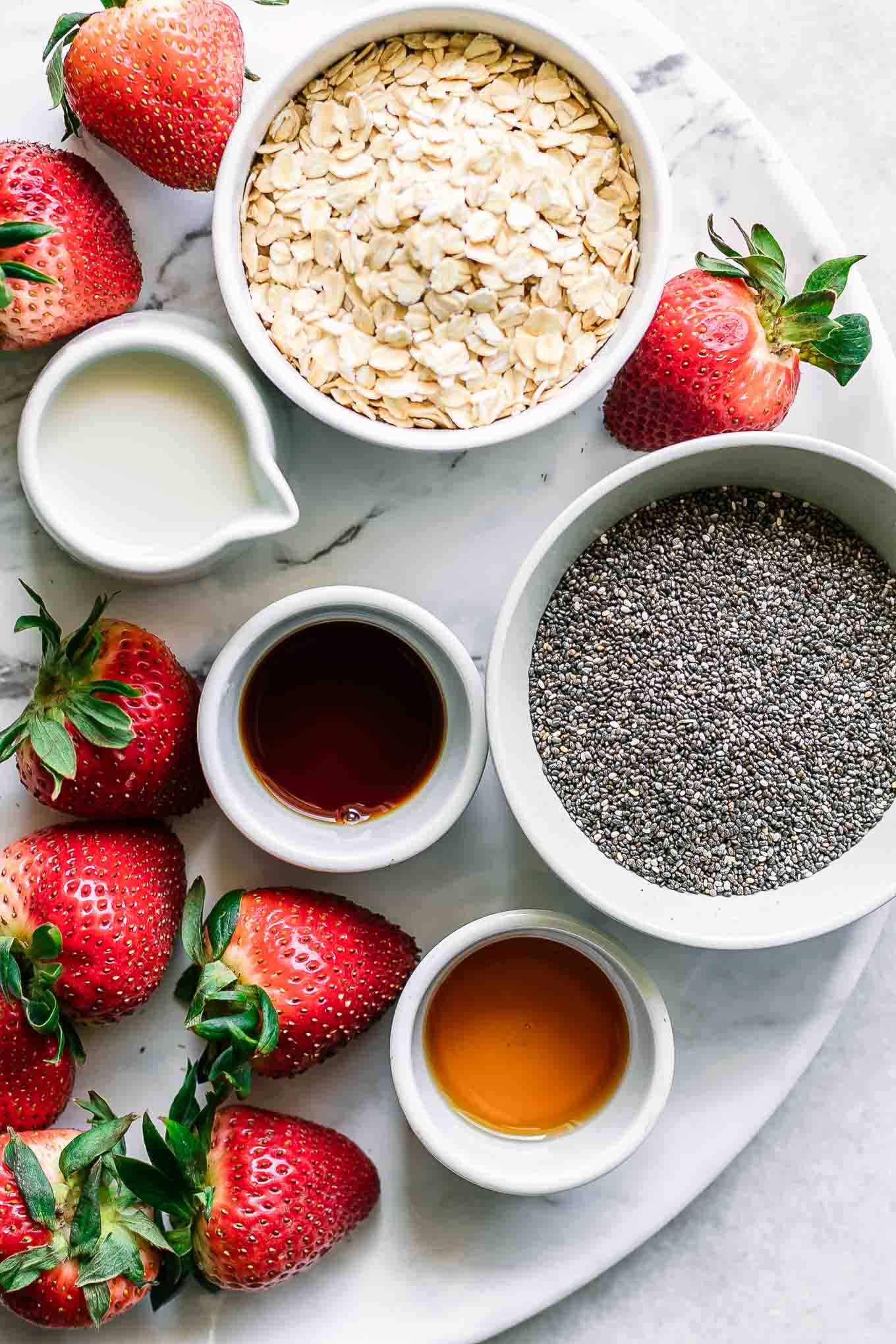 bowls of rolled oats, chia seeds, milk, maple syrup, vanilla, and strawberries on a white table for overnight oats