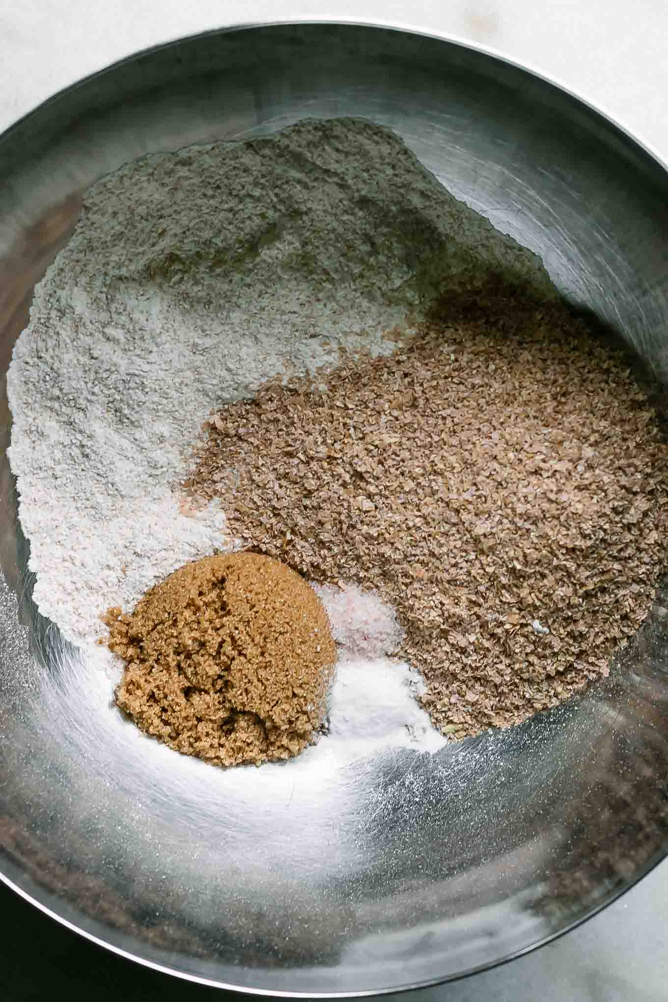 a mixing bowl with wheat bran, wheat flour, brown sugar, and other dry muffin ingredients on a white table