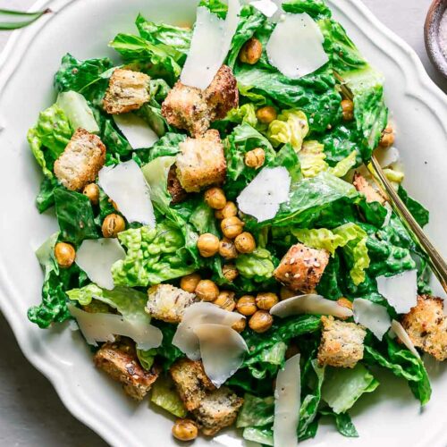 a white plate with vegan caesar salad with croutons, chickpeas, and shaved vegan parmesan cheese on a white table