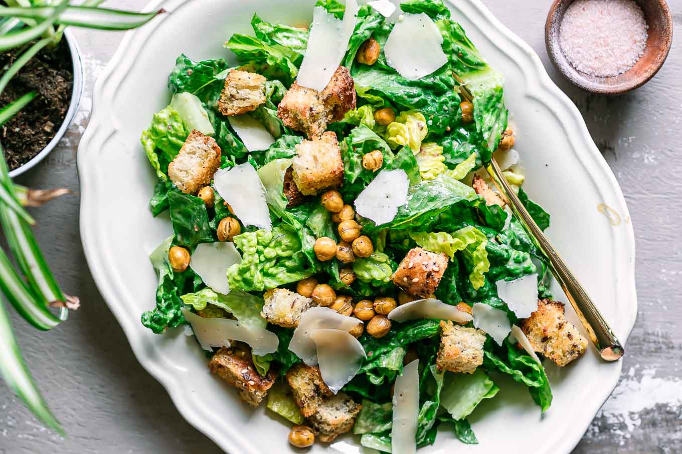 a white plate with vegan caesar salad with croutons, chickpeas, and shaved vegan parmesan cheese on a white table