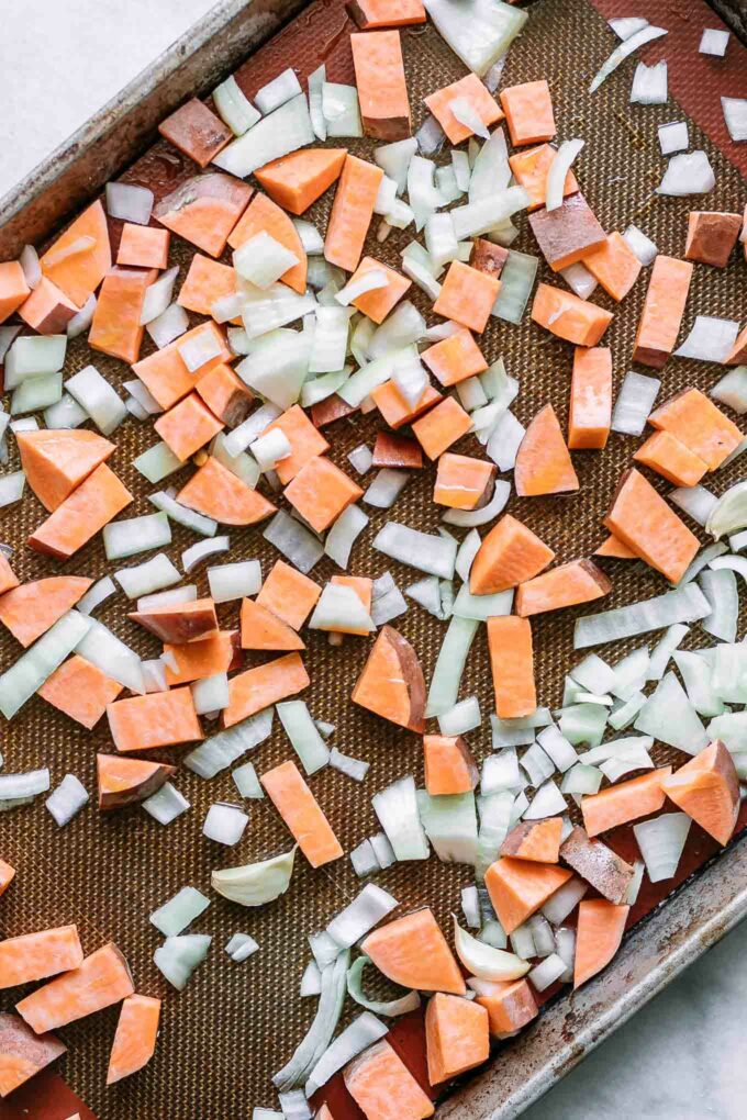 a baking sheet with cut sweet potatoes, yellow onion, and garlic before baking