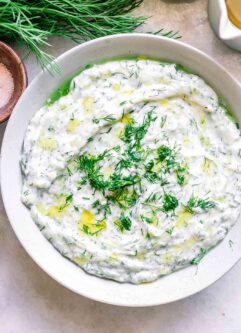 a white bowl with vegan tzatziki sauce with fresh dill and a lemon on a white table