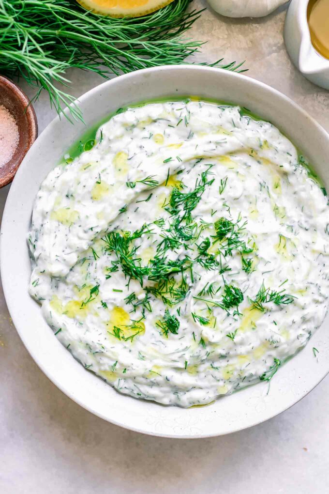 a white bowl with vegan tzatziki sauce with fresh dill and a lemon on a white table
