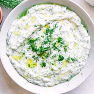 a white bowl with vegan tzatziki sauce on a table with fresh dill and lemons