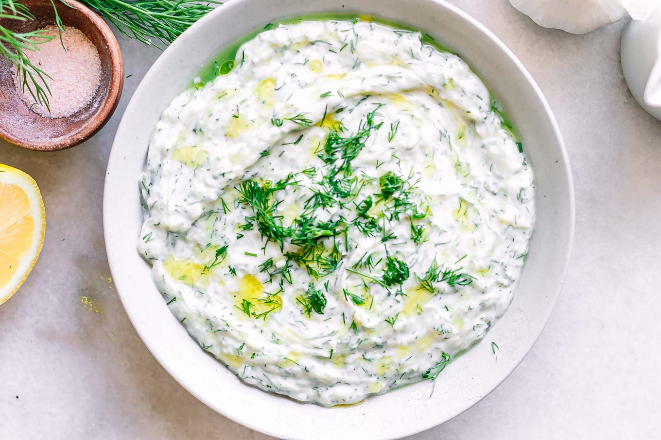 a white bowl with vegan tzatziki sauce on a table with fresh dill and lemons
