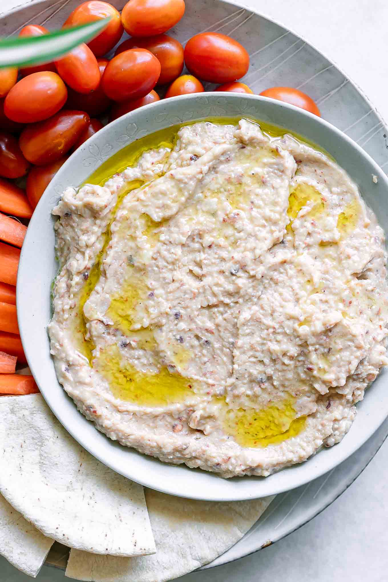 a bowl of black eyed pea hummus with pita bread, carrot sticks, and cherry tomatoes on a white table