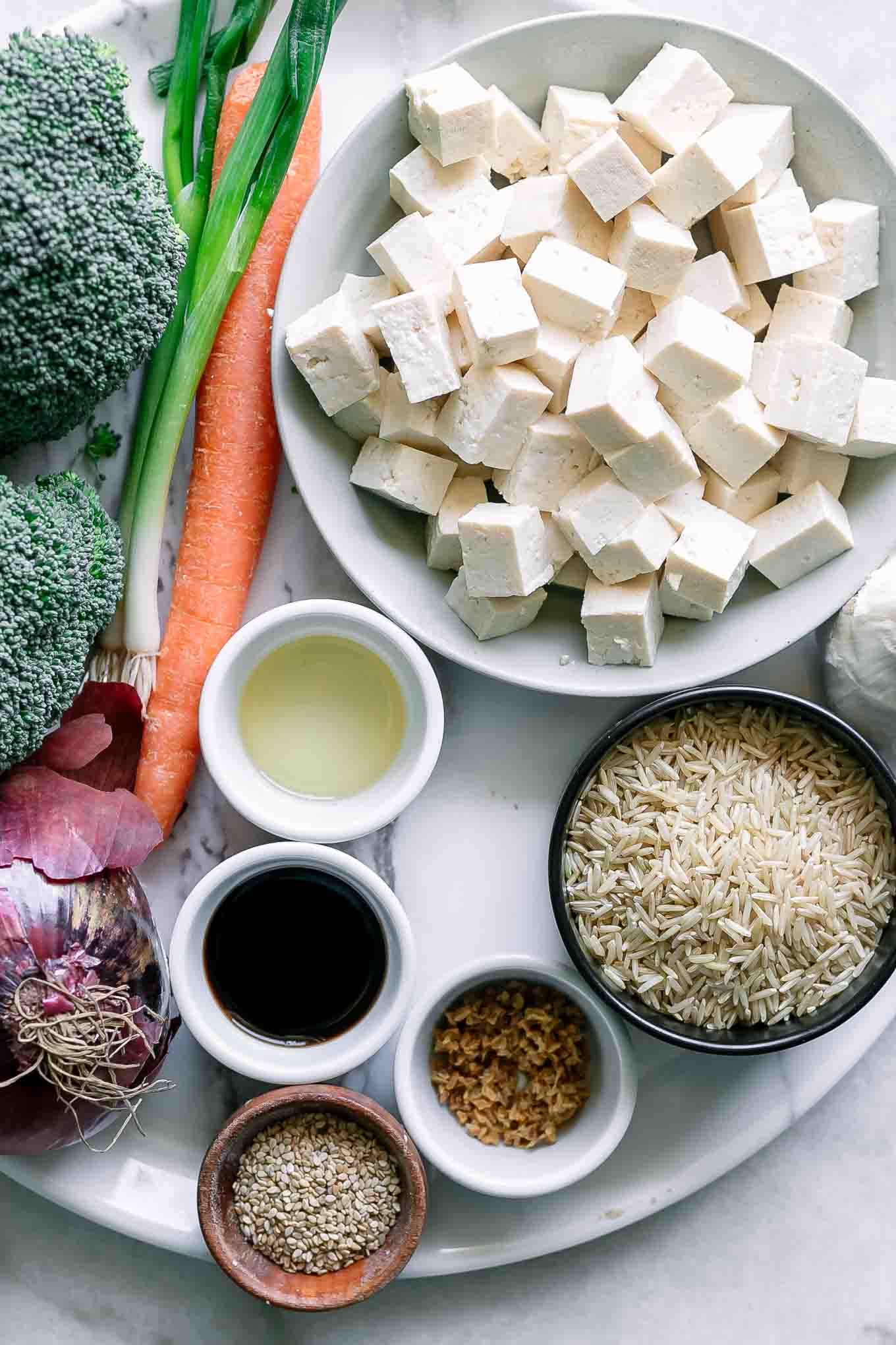 bowls of tofu, rice, broccoli, carrots, onion, and spices on a table before cooking