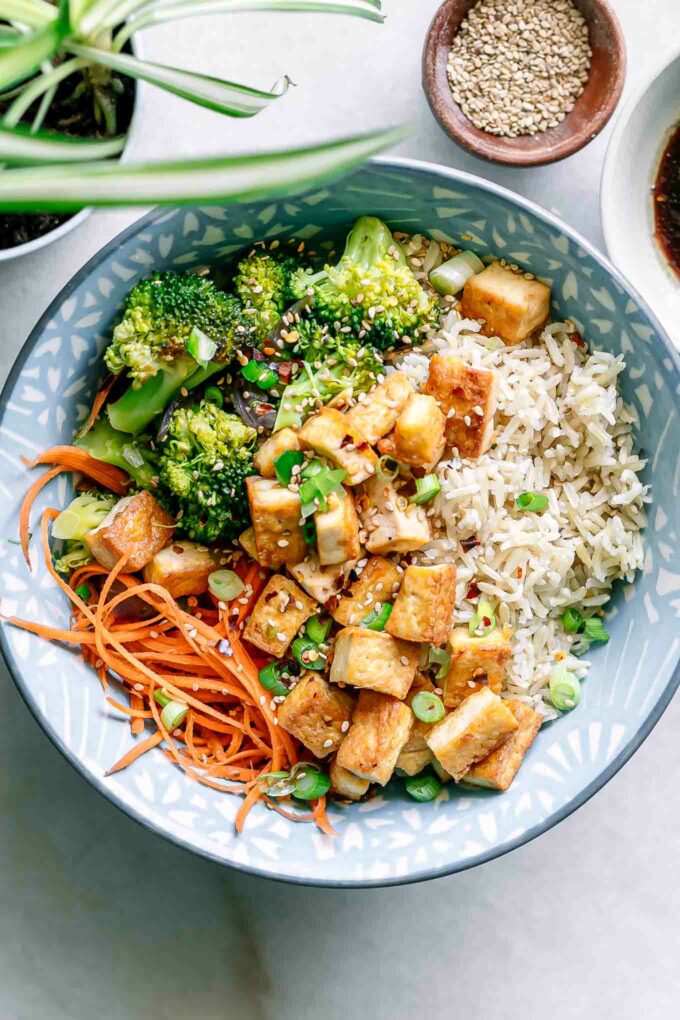 a blue bowl with brown rice, tofu, broccoli, shredded carrots, and sesame seeds on a white table