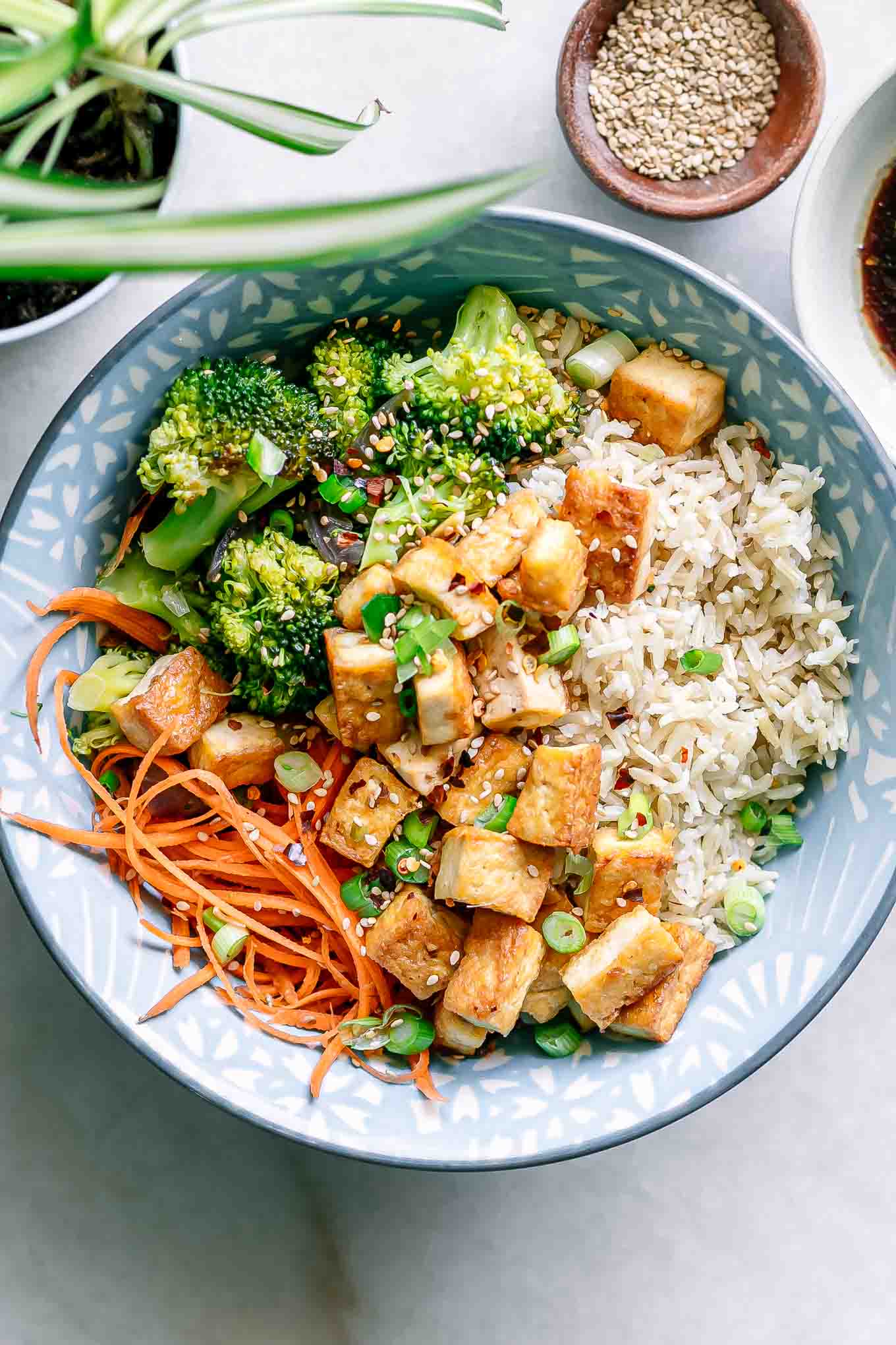 a blue bowl with brown rice, tofu, broccoli, shredded carrots, and sesame seeds on a white table