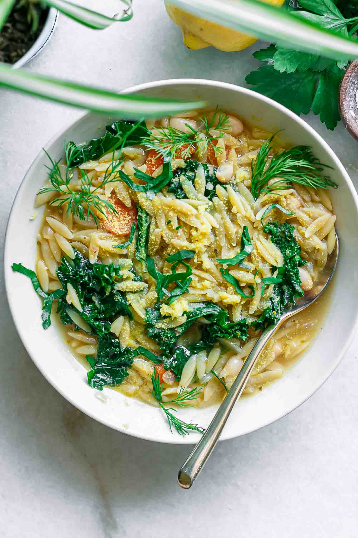 a white bowl of kale lemon white bean orzo soup with a gold fork on a white table