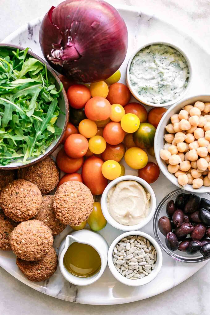 arugula, onion, tomato, chickpeas, olives, hummus, tzatziki, seeds, and falafel on a white table for a salad