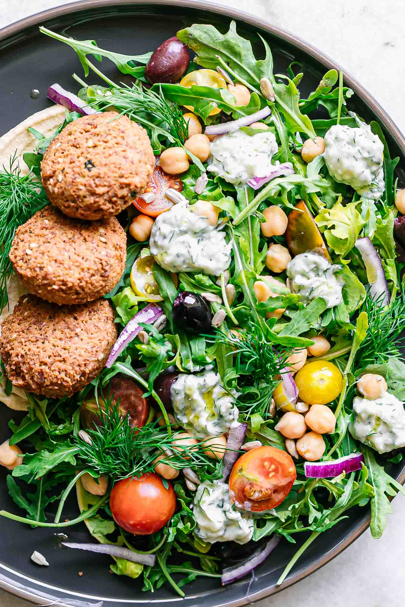 a plate with arugula salad with falafel, red onion, olives, tomatoes, tzatziki, hummus, and sunflower seeds