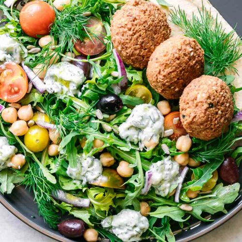 an Mediterranean-style salad with arugula, falafel, chickpea, tomatoes, onion, hummus, tzatziki, and sunflower seeds on a blue plate