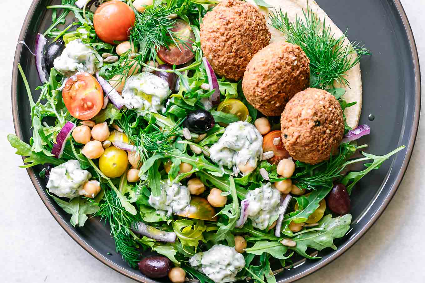 an Mediterranean-style salad with arugula, falafel, chickpea, tomatoes, onion, hummus, tzatziki, and sunflower seeds on a blue plate