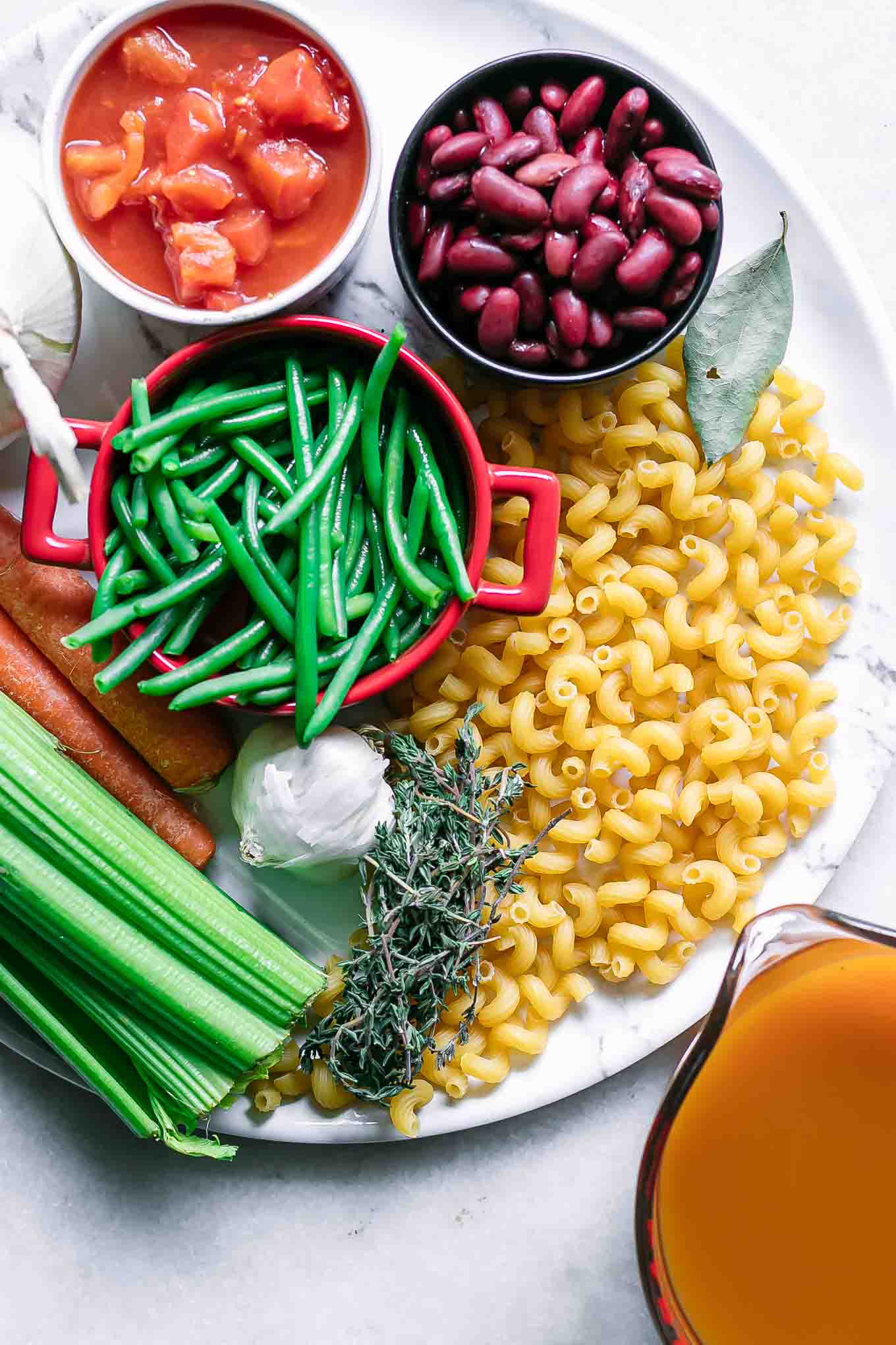 bowls of tomatoes, beans, green beans, pasta, garlic, spices, and vegetables on a table for minestrone soup