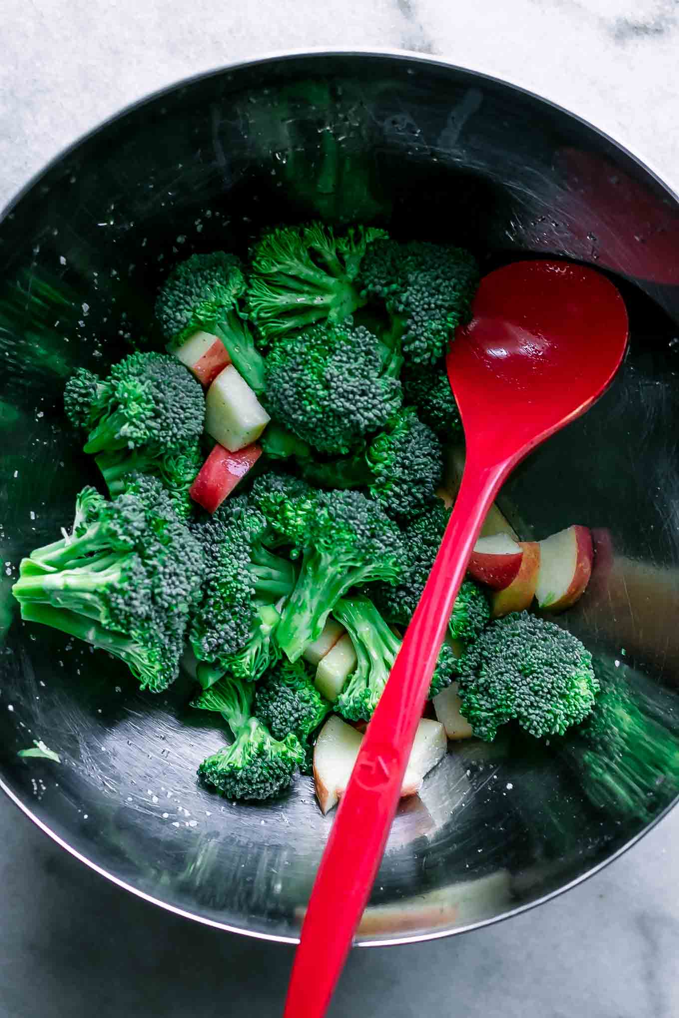a mixing bowl with cut broccoli florets and sliced apples with a red spoon