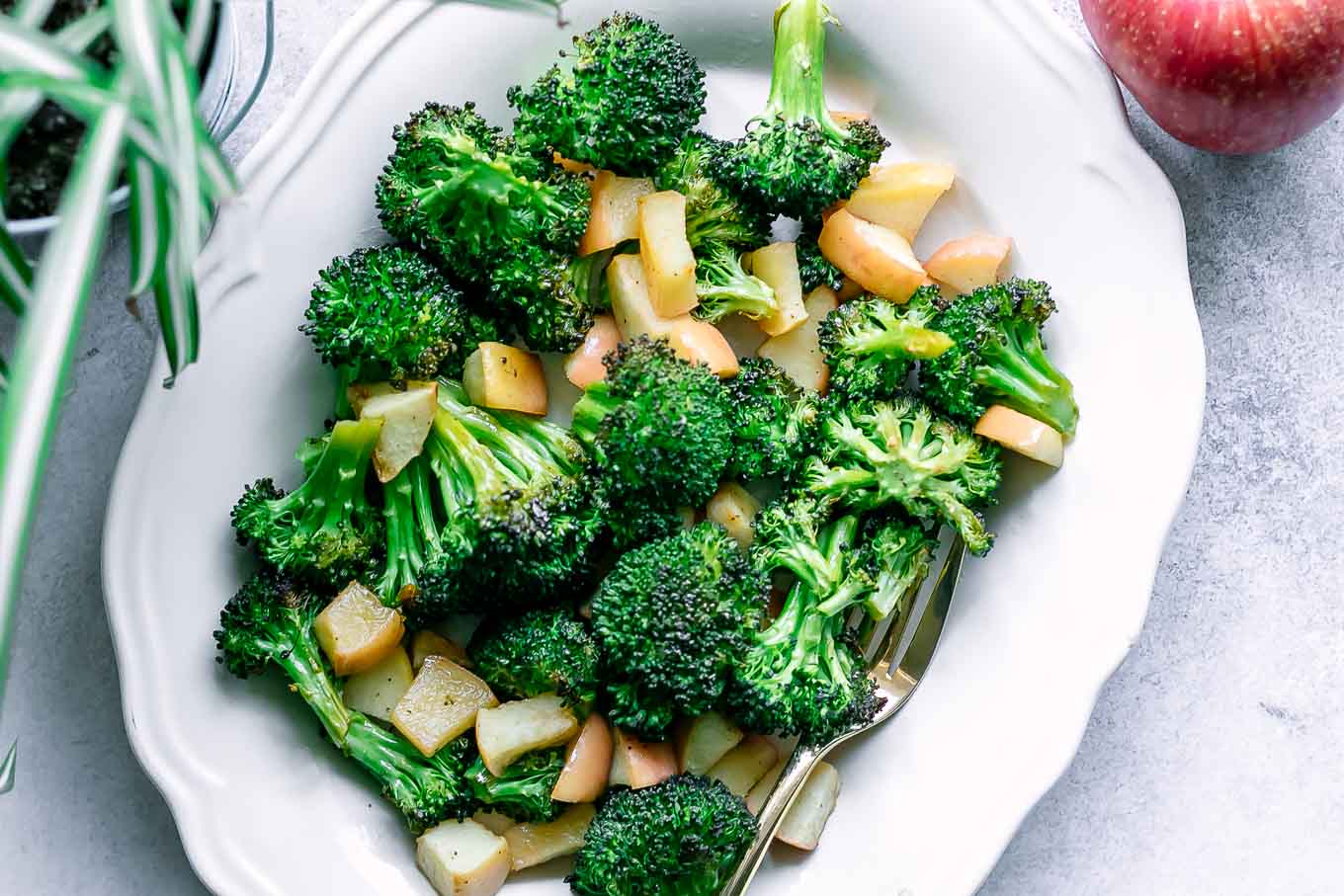 a white plate with baked broccoli and apples side dish on a white table