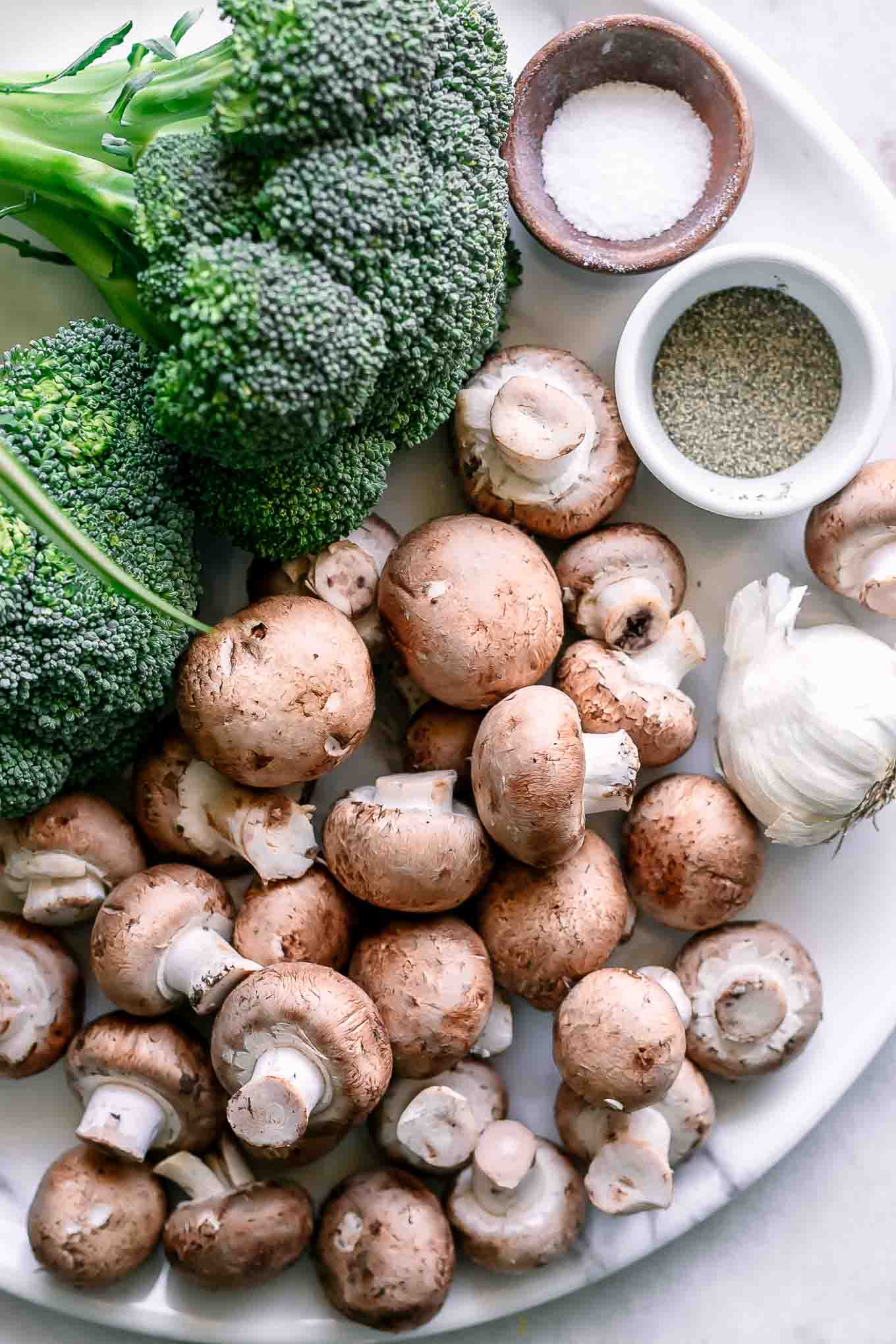 broccoli, mushrooms, garlic, and bowls of spices on a table before roasting