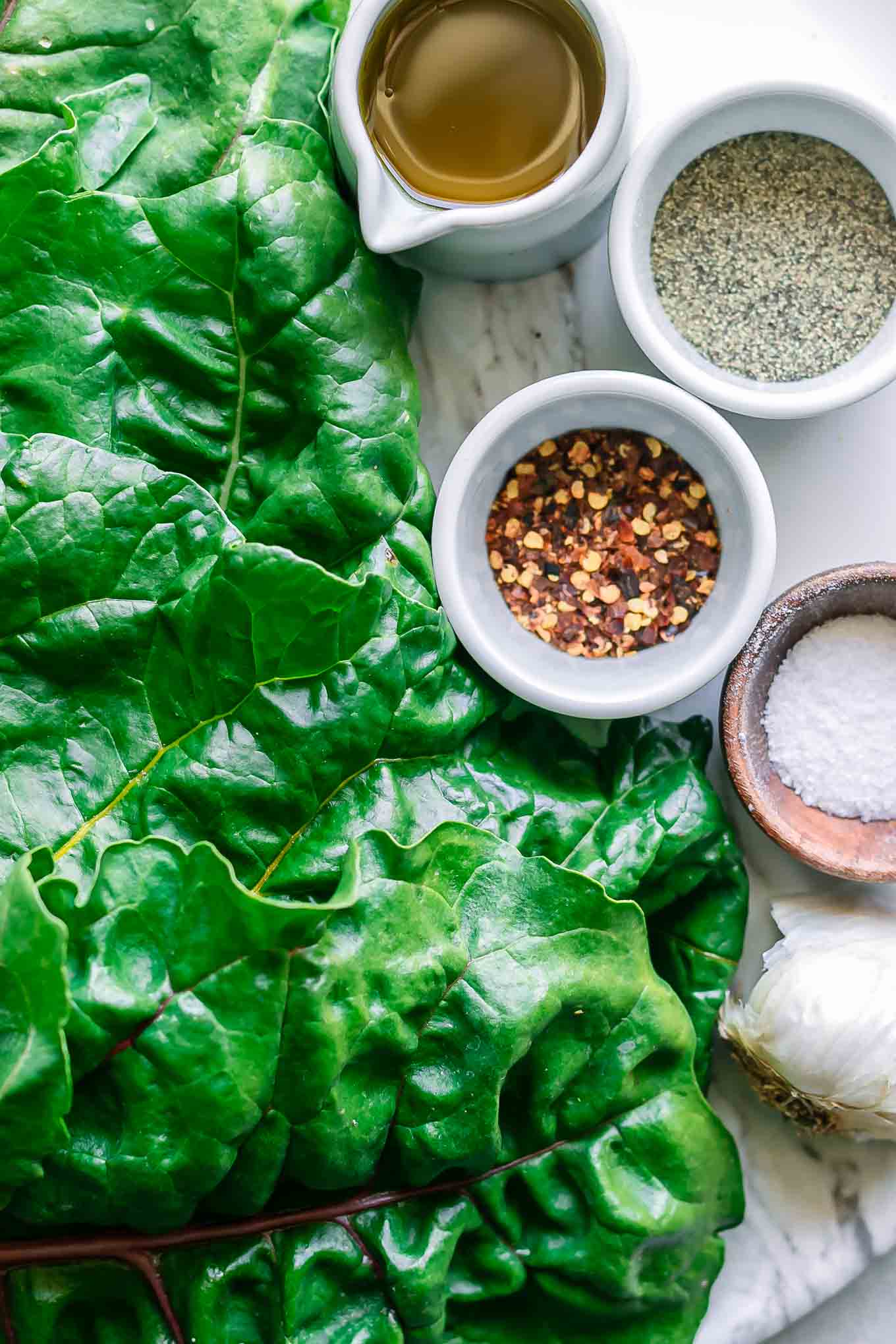 swiss chard leaves on a table with bowls of oil, garlic, salt, and pepper for sautéing