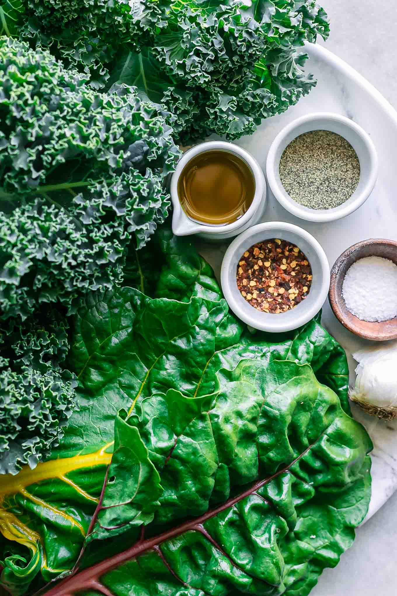 fresh kale and chard leaves with bowls of olive oil, garlic, salt, pepper, and red pepper flakes