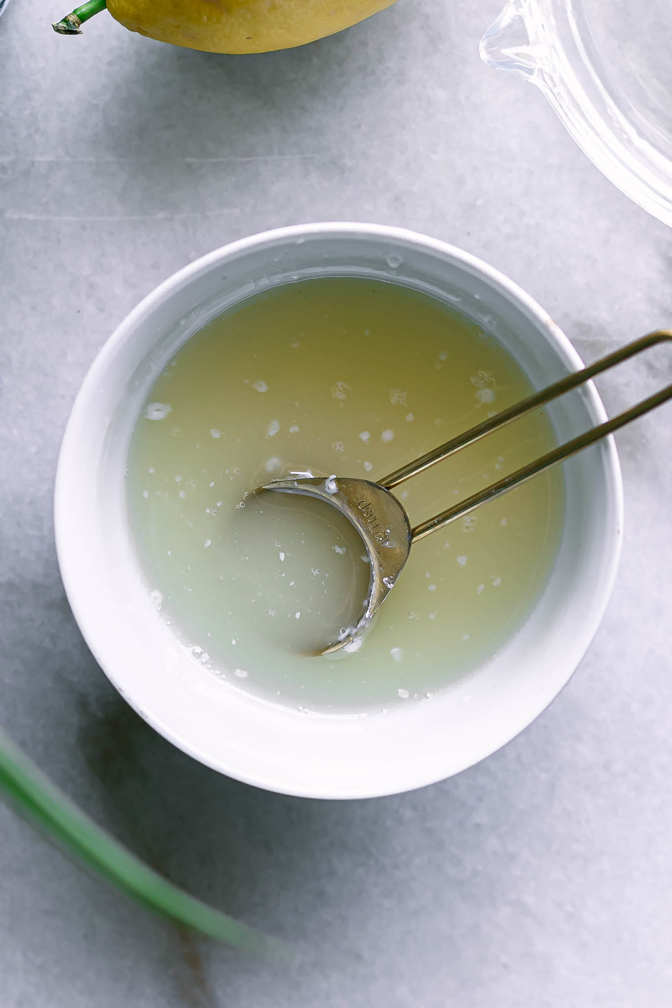 a bowl of lemon glaze sauce with a gold spoon on a white table with a fresh lemon