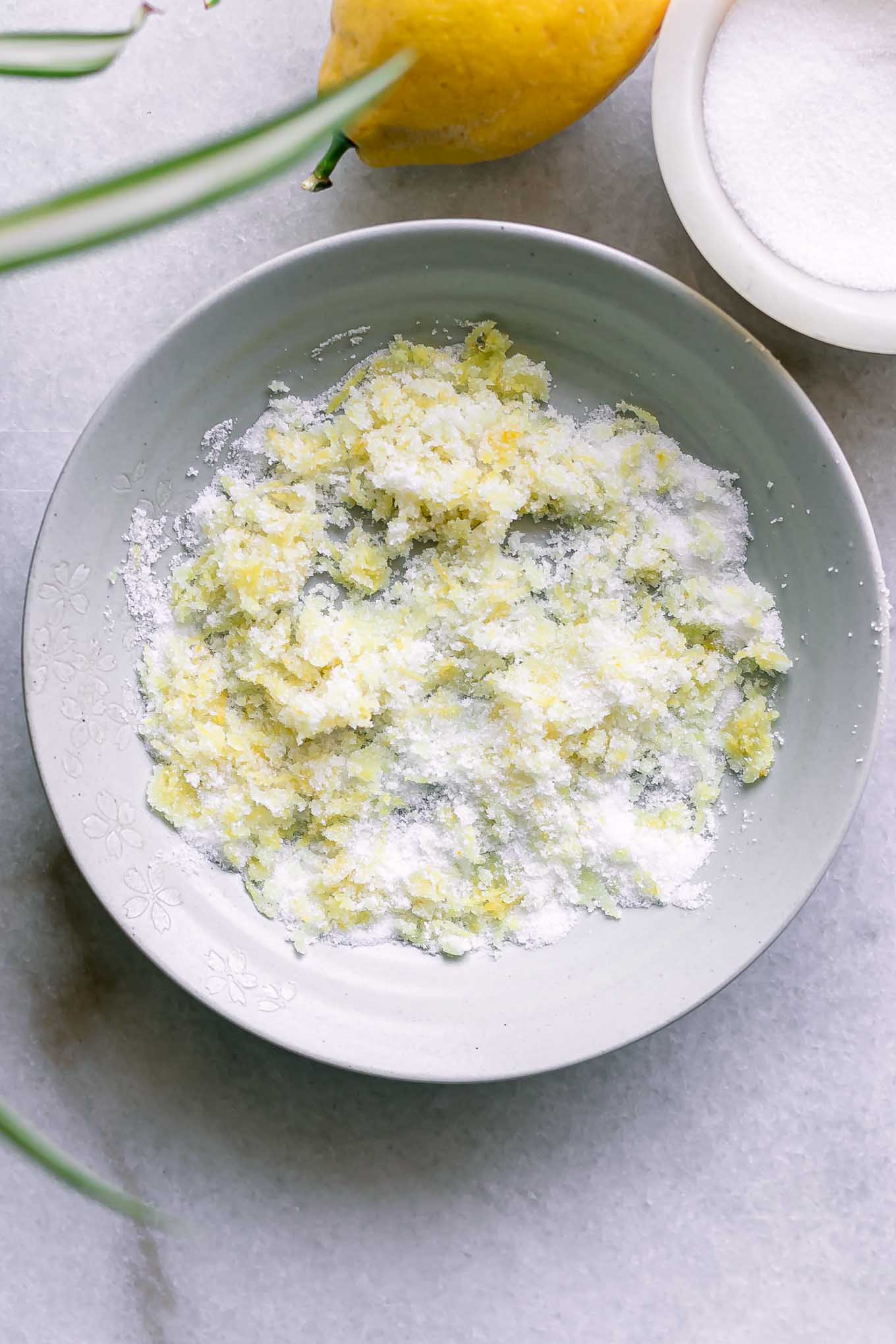 a bowl of white sugar with lemon zest inside on a table with a lemon