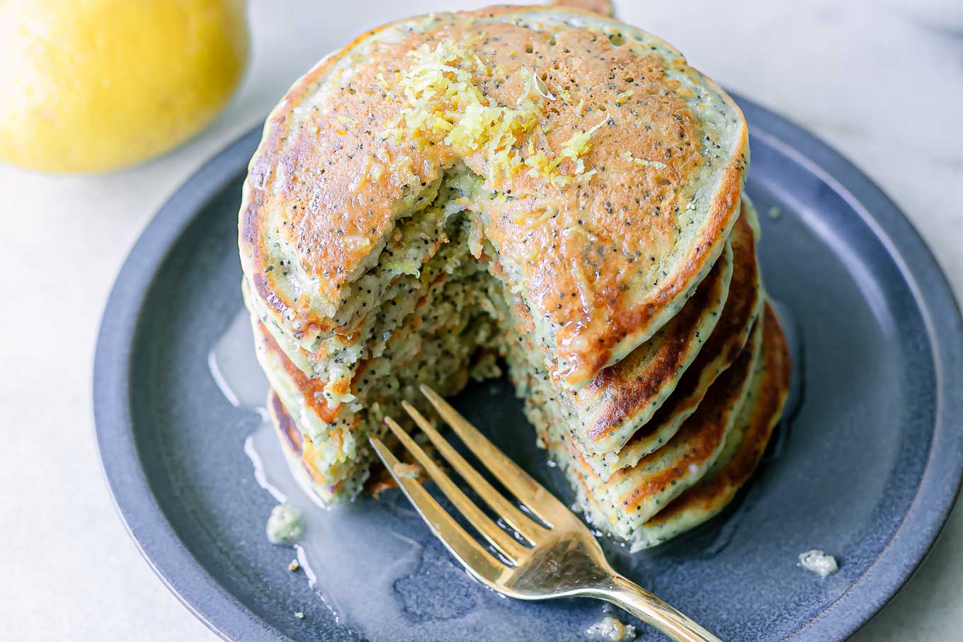 a blue plate with a stack of vegan lemon poppyseed pancakes with a gold fork and a whole lemon