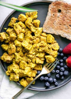 a blue plate with a yellow tofu scramble, a piece of bread, and a berries on a white table