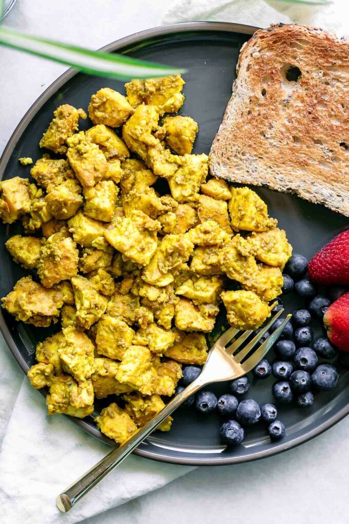 a blue plate with a yellow tofu scramble, a piece of bread, and a berries on a white table