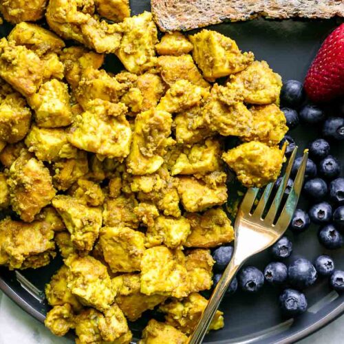 a blue plate with a yellow tofu scramble with fresh berries and a gold fork on a white table