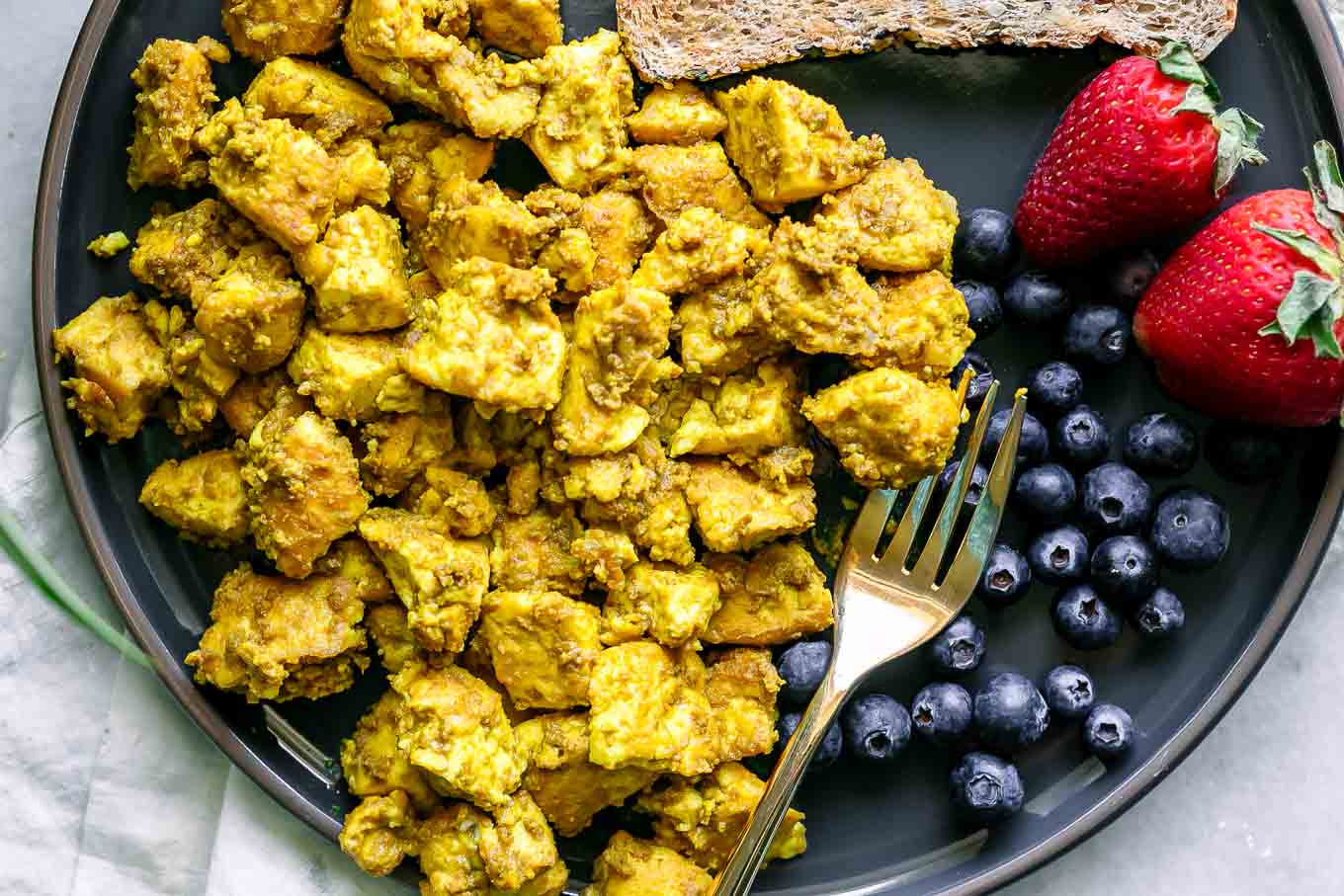 a blue plate with a yellow tofu scramble with fresh berries and a gold fork on a white table
