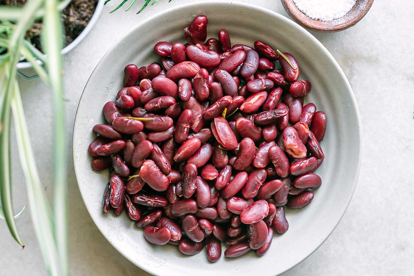 a bowl of cooked kidney beans on a white table with herbs and a bowl of salt