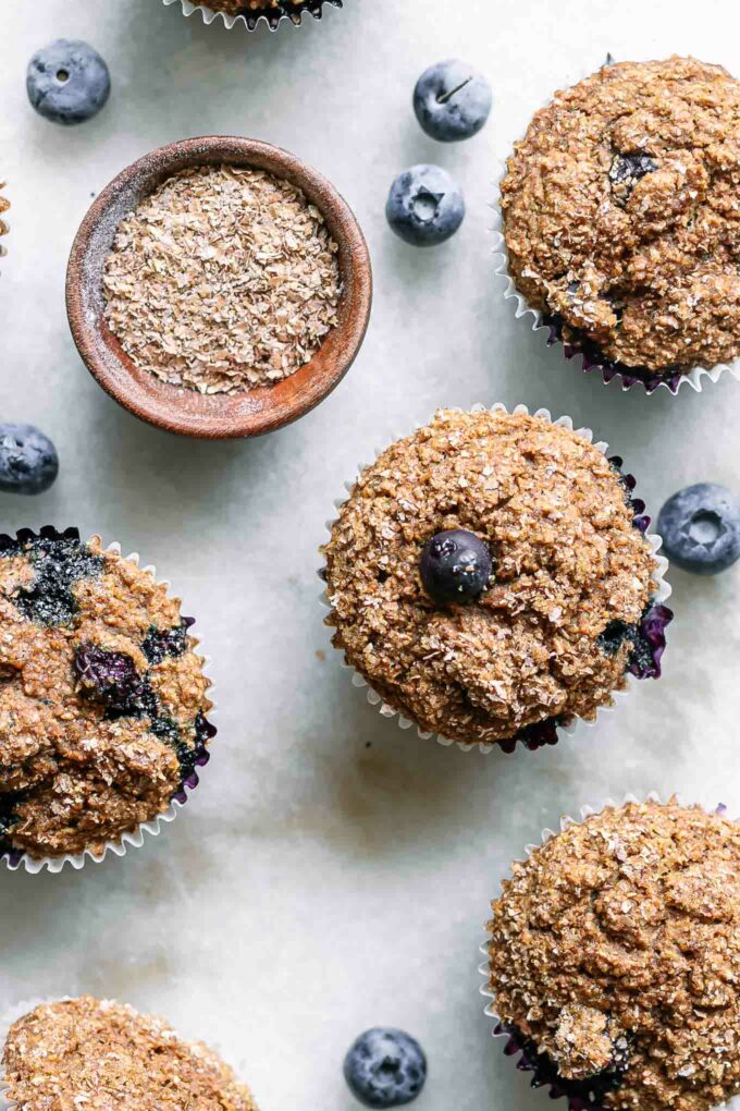plant-based blueberry bran muffins on a white table with a bowl of wheat bran and fresh blueberries