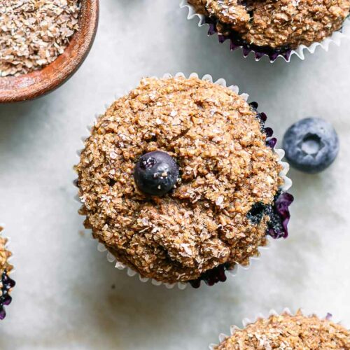plant-based blueberry bran muffins on a white table with fresh blueberries and a bowl of wheat bran
