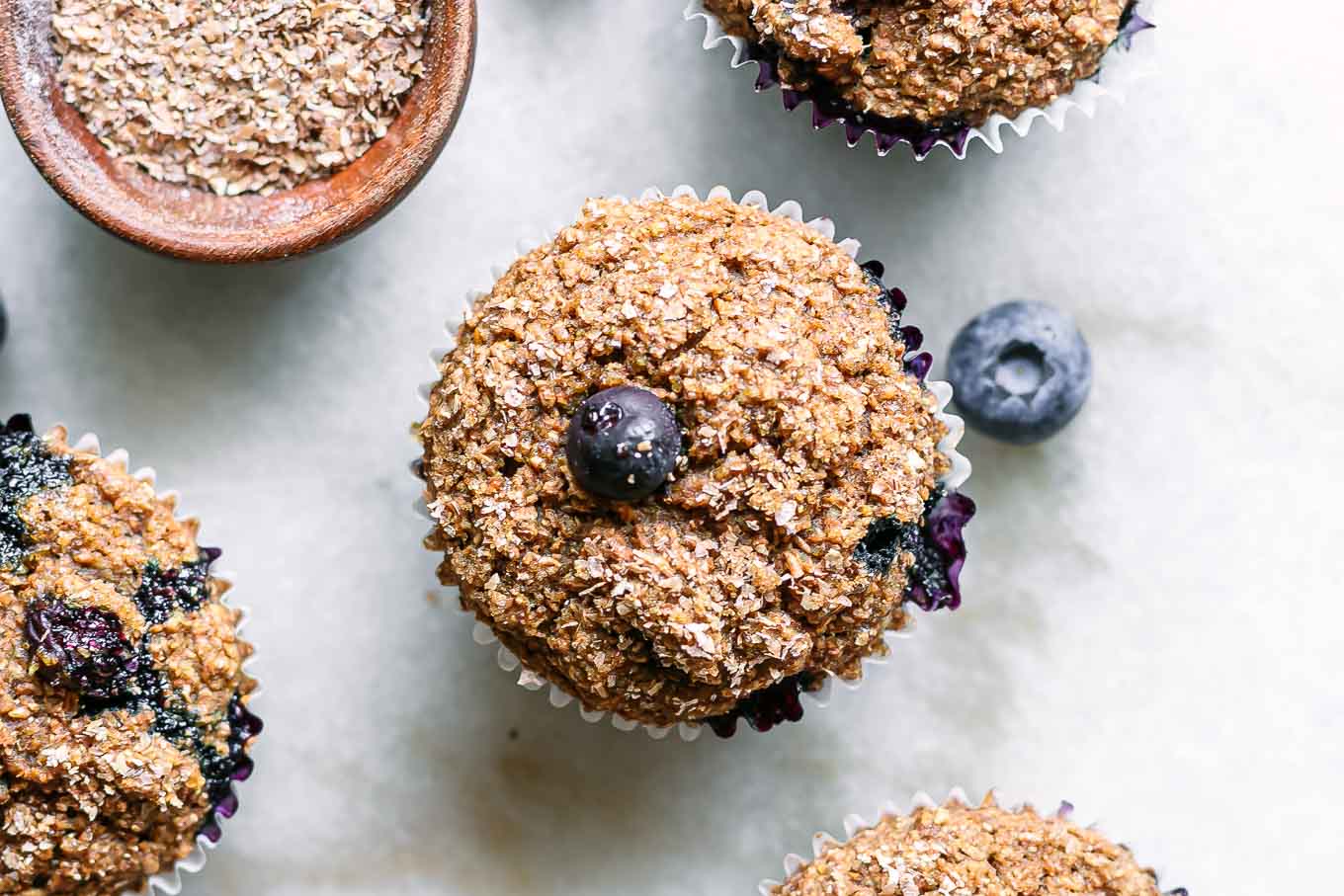 plant-based blueberry bran muffins on a white table with fresh blueberries and a bowl of wheat bran