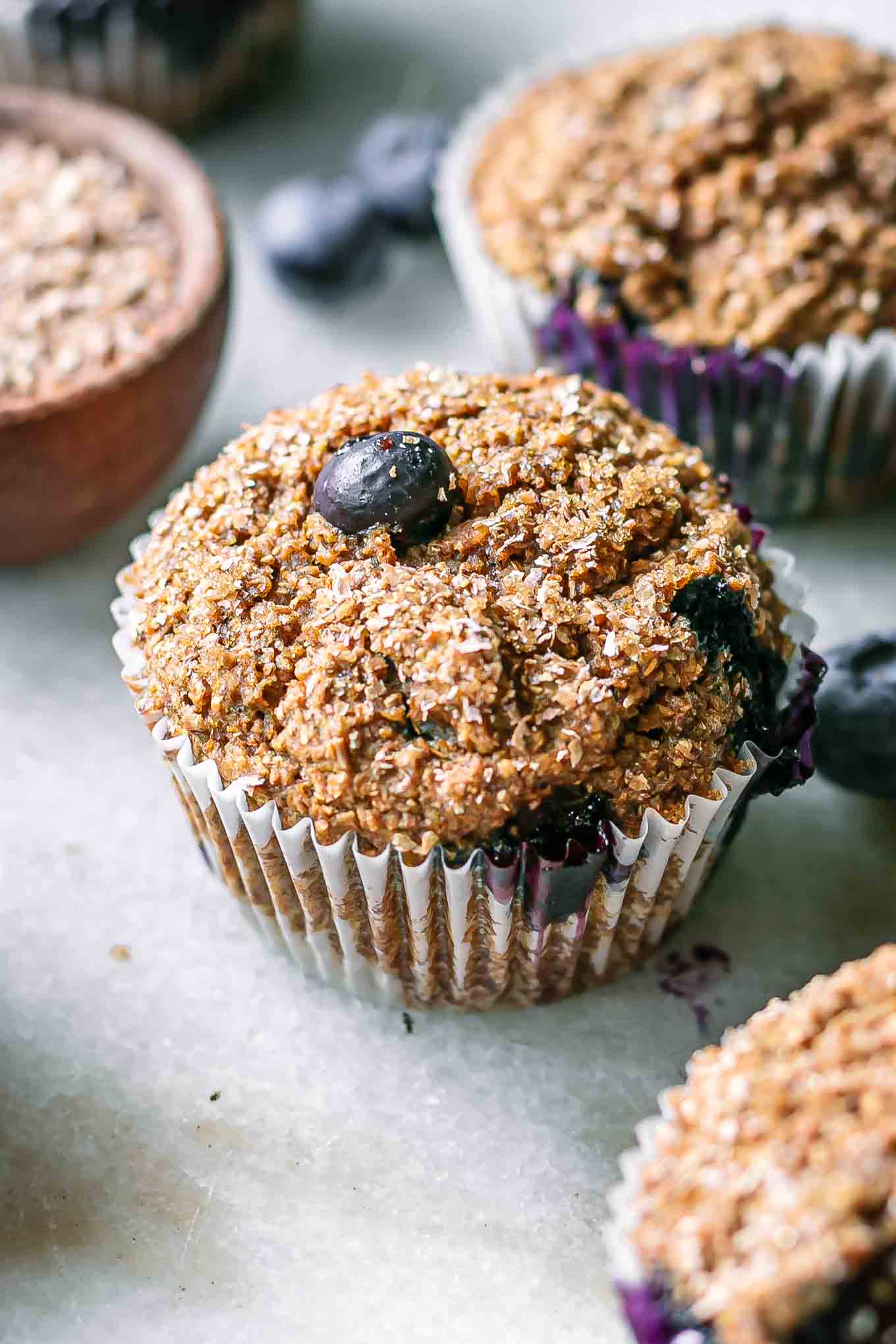 a close up photo of a vegan blueberry bran muffin on a white table with fresh blueberries