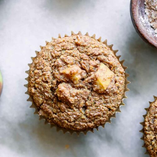 an apple bran muffin on a table with a whole apple and a bowl of wheat bran