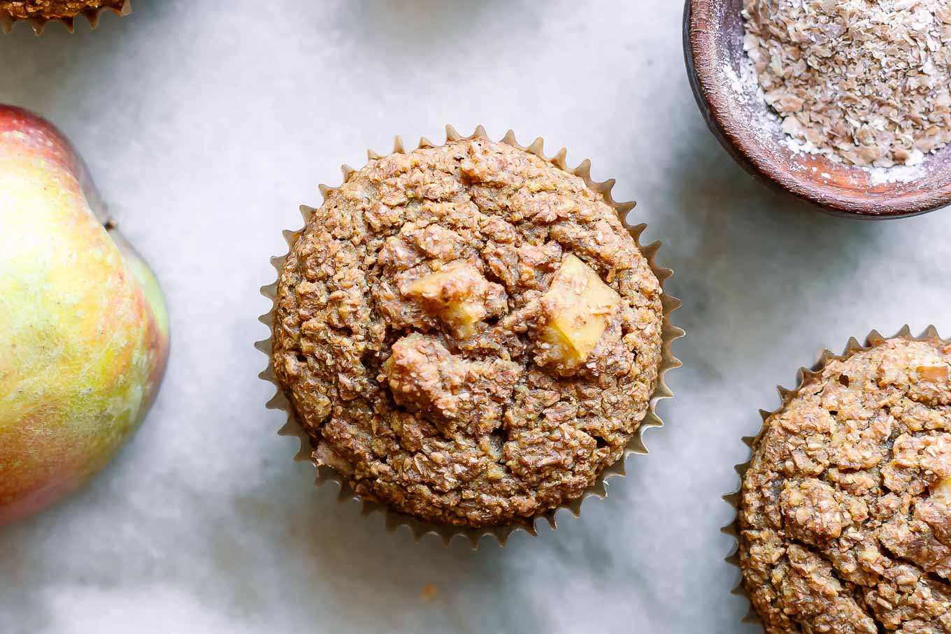 an apple bran muffin on a table with a whole apple and a bowl of wheat bran