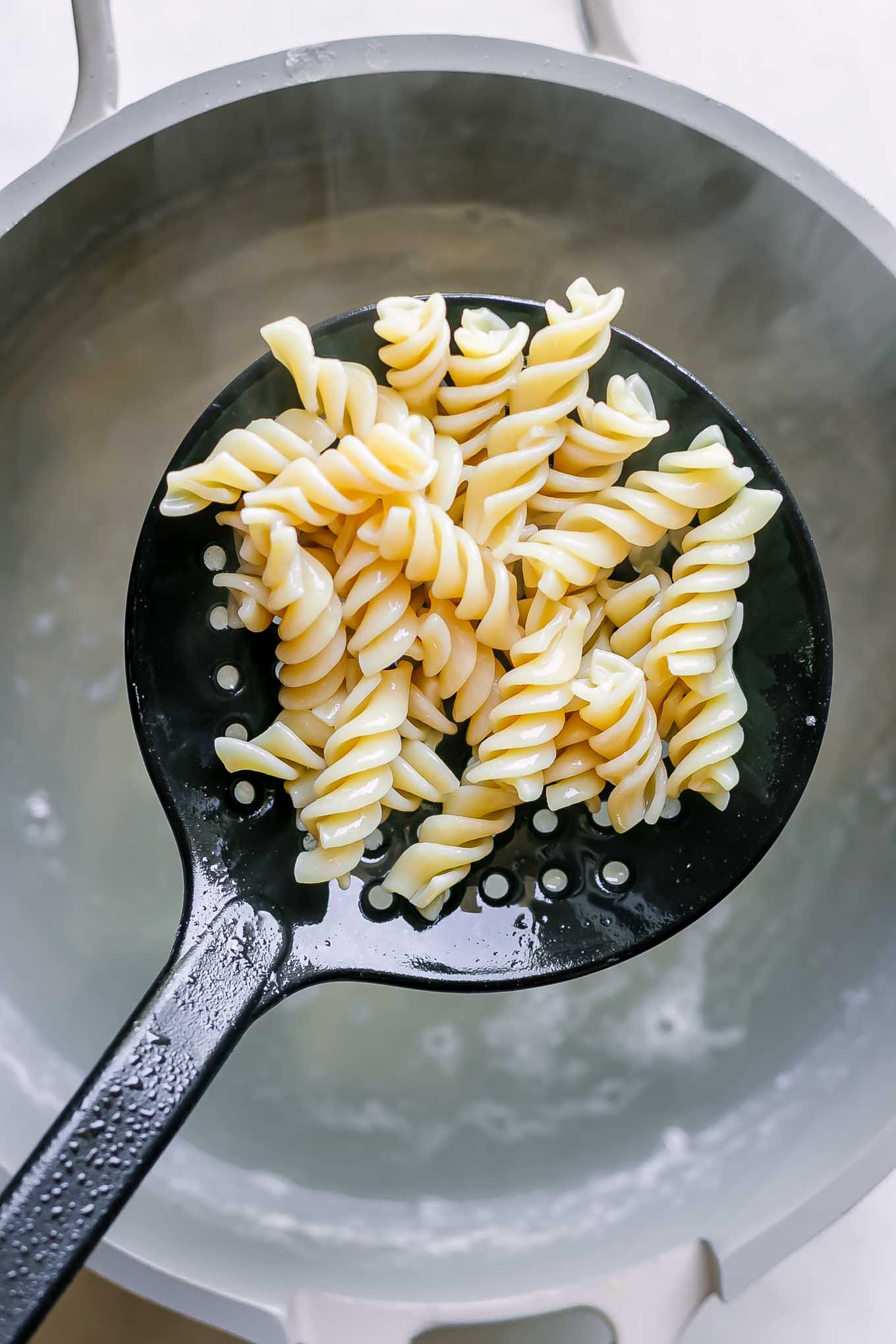 a large black spoon lifting cooked pasta out of a pot of boiling water
