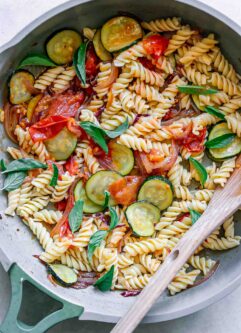 a pan of rotini pasta with zucchini, tomatoes, and basil on a white table