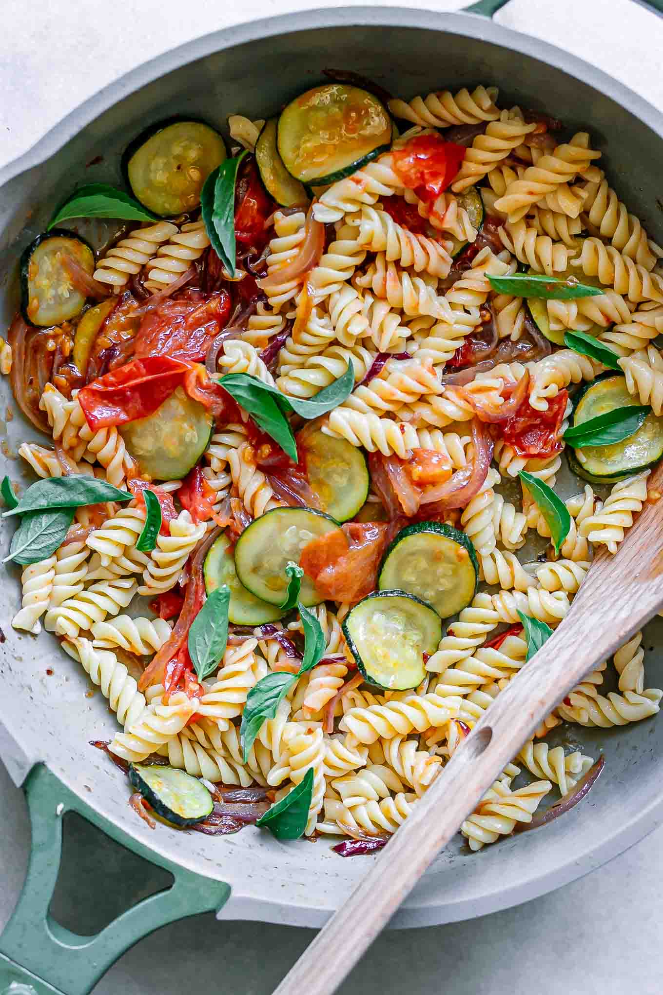 a pan of rotini pasta with zucchini, tomatoes, and basil on a white table