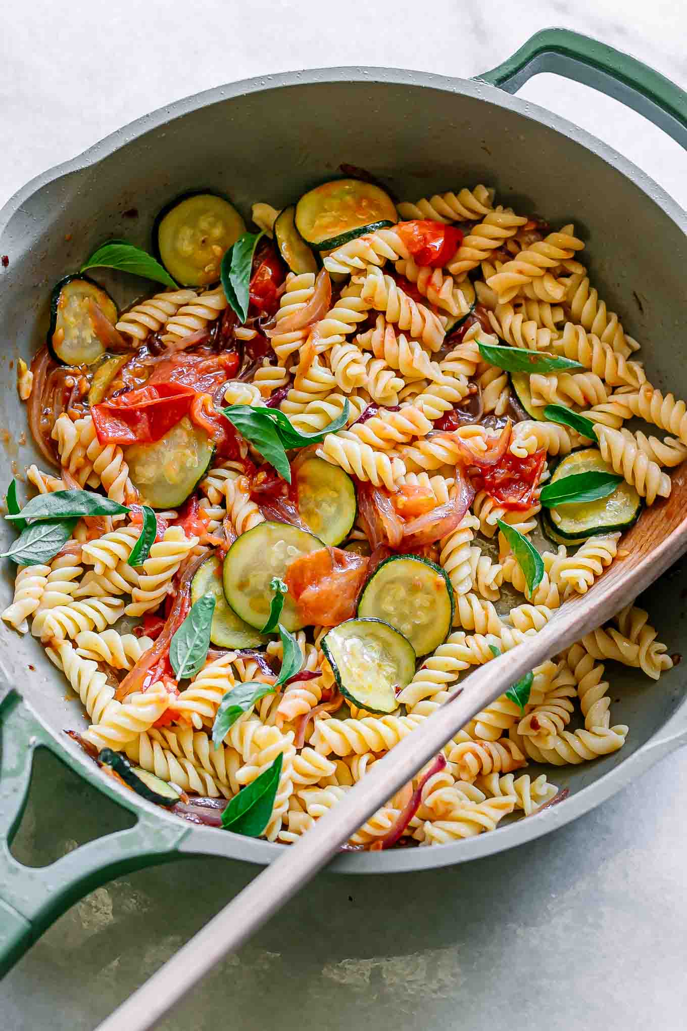 zucchini and tomato pasta in a pan with a wooden spoon on a white table