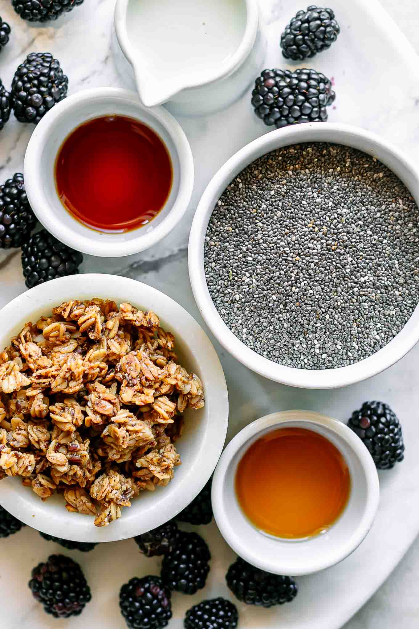 bowls of chia seeds, blackberries, milk, vanilla, maple syrup, and granola on a white table