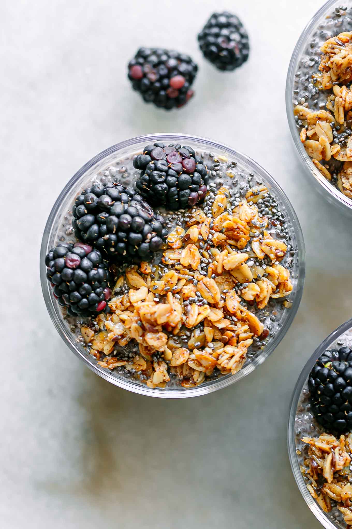 a top down photo of the top of three glasses of blackberry chia pudding with granola and fresh blackberries as garnish