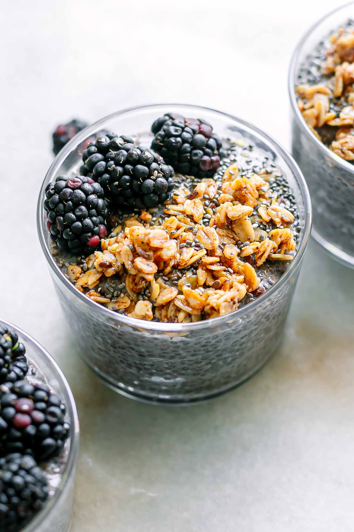 three jars of purple chia seed pudding with blackberries and granola on a white table