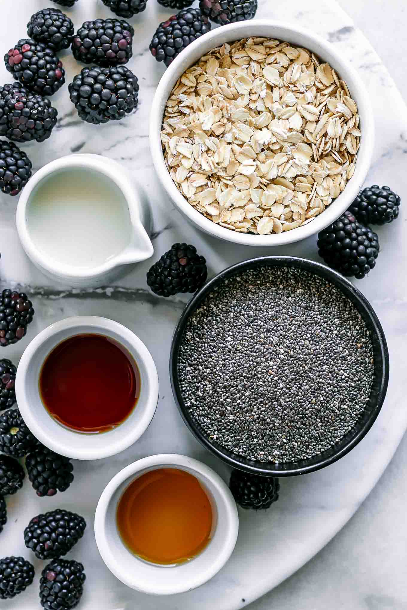 bowls of oats, chia seeds, milk, vanilla, maple syrup and blackberries on a white table