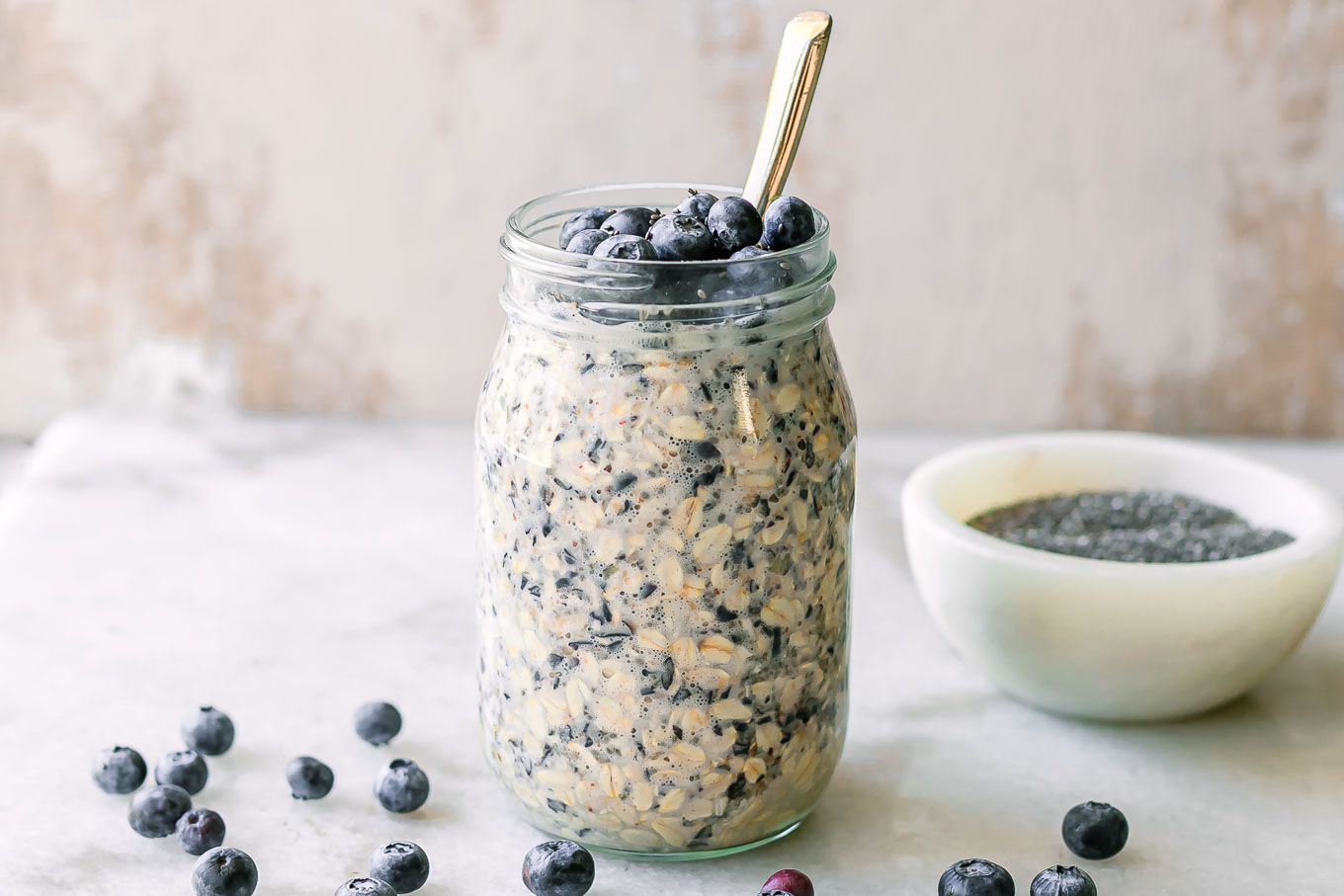 a jar of blueberry overnight oats with fresh blueberry garnish on a white table with a gold spoon
