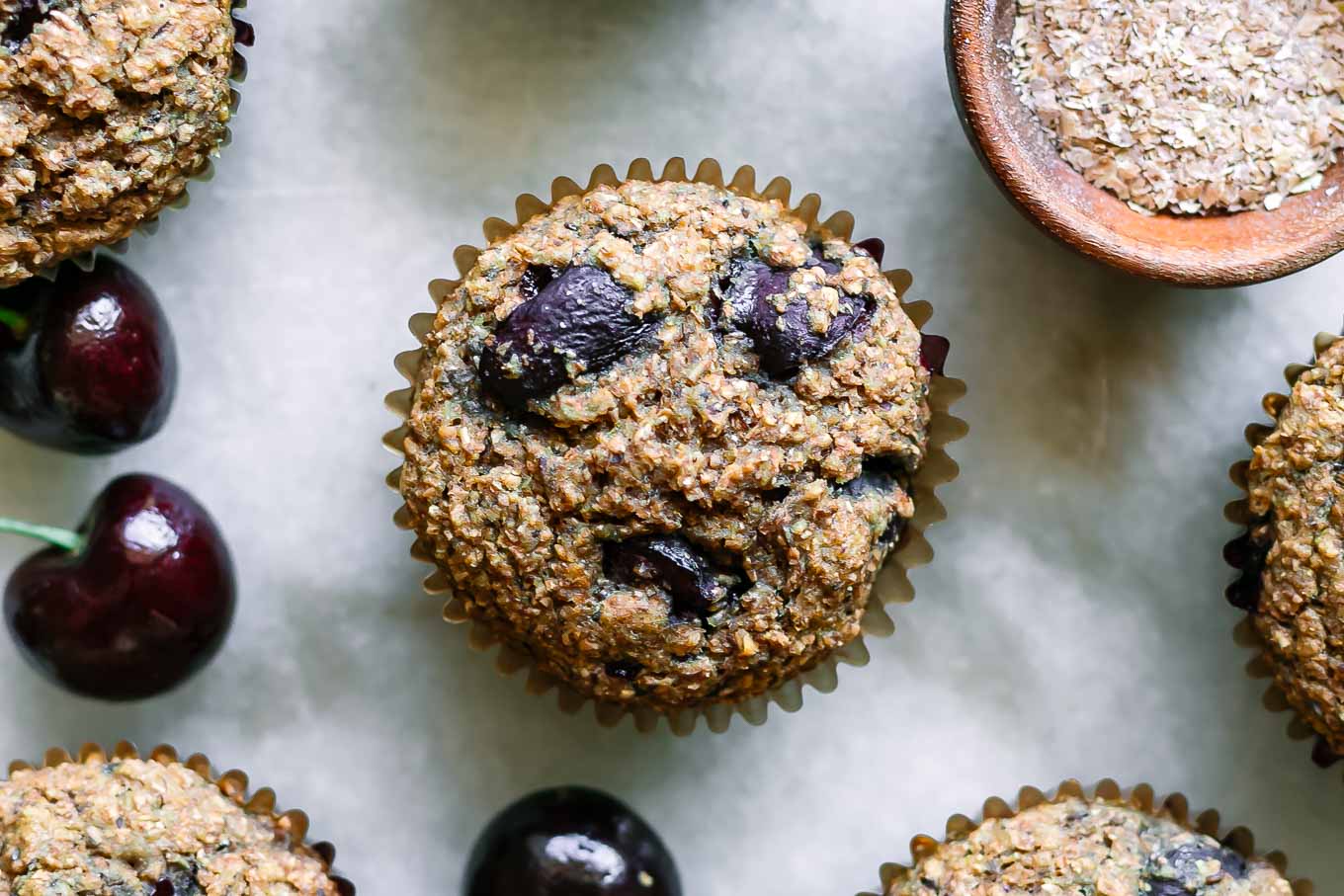 cherry bran muffins on a white table with fresh cherries and a bowl of wheat bran