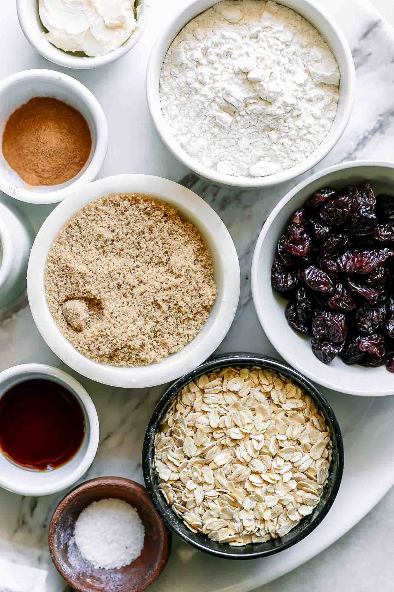 bowls of oatmeal, flour, brown sugar, dried cherries, and other ingredients for cookies