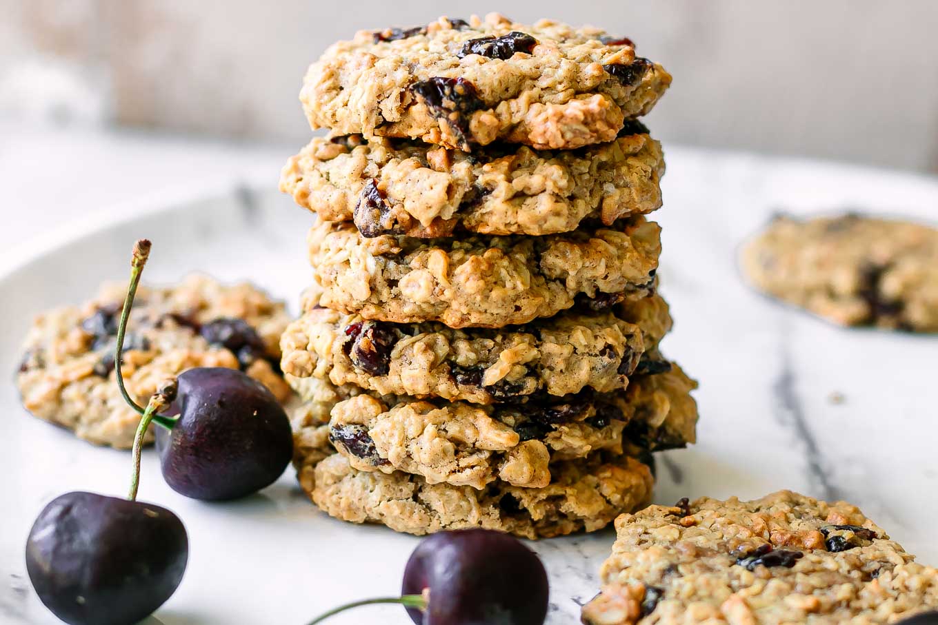 stacked plant-based cherry oatmeal cookies on a white table with fresh black cherries